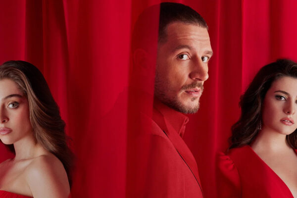 Three models in red outfits pose behind red curtains, facing the camera with neutral expressions (promotional fashion photo).