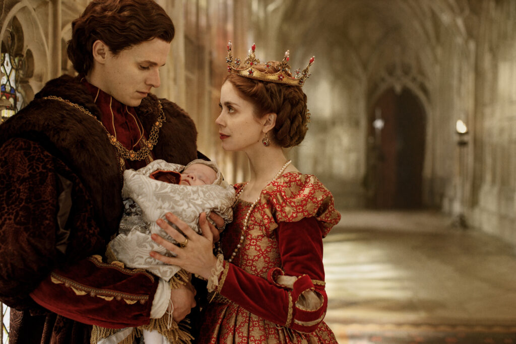The Spanish Princess Season 2 2020 - viasatepicdrama.com A royal couple in ornate medieval gowns cradle a swaddled baby, standing in a stone hallway with warm lighting and a crown-wearing mother looking at the infant.